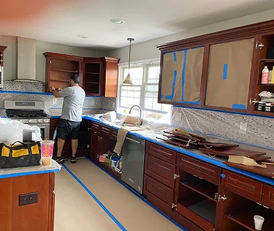 Man working on kitchen cabinets during renovation, with blue tape and protective coverings.