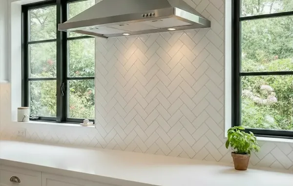 Kitchen backsplash with white subway tile in a herringbone pattern, stainless steel range hood, and two windows overlooking greenery.